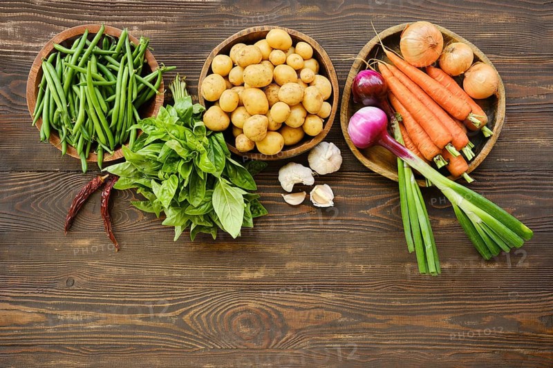 Bunches of green beans, baby potatoes and carrots are placed on wooden table. Garlic, green onions, and herbs are also visible. All items are fresh and suitable for meal preparation