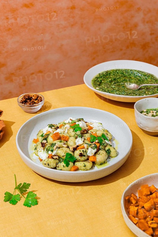 White plate filled with delicious Italian gnocchi mixed with bright green pesto, diced vegetables, walnuts, and crumbled cheese, beautifully arranged against a vibrant orange background