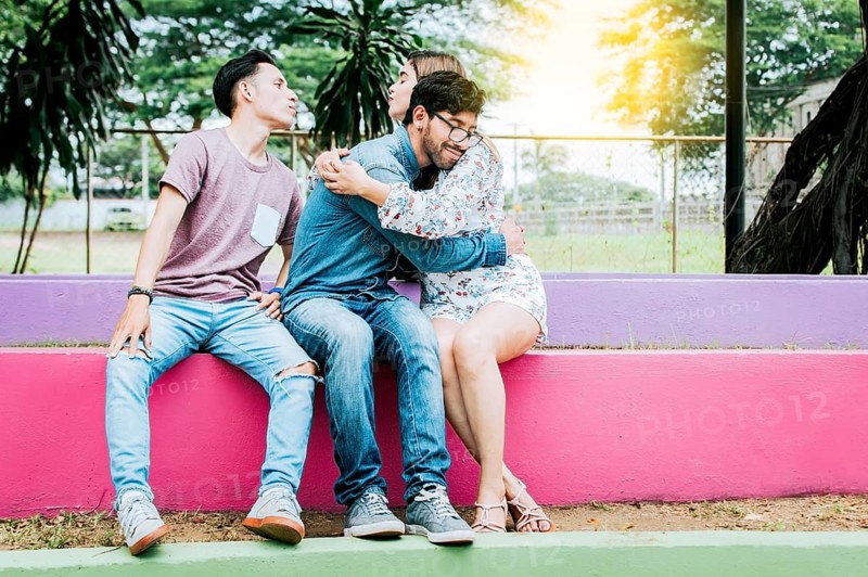 Unfaithful girl sitting hugging her boyfriend and secretly kissing another man. Unfaithful girlfriend hugging her boyfriend in a park secretly kissing another man, Couple infidelity concept
