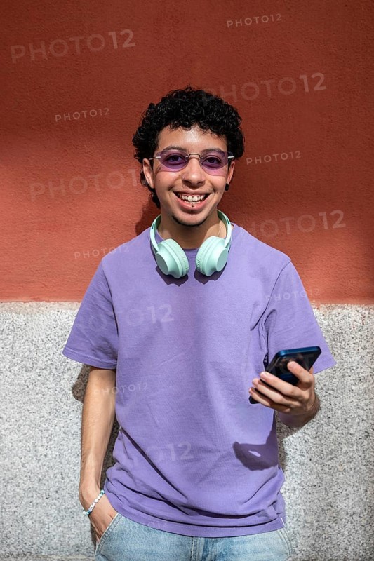A LGBTQ+ cheerful person stands against a wall, wearing headphones and holding a phone. They exude a relaxed and confident vibe, dressed in a purple shirt and sunglasses