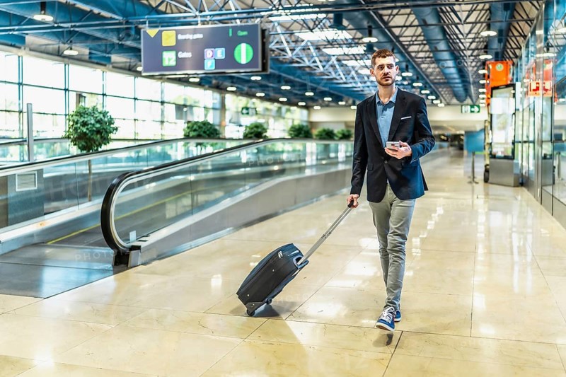 Businessman pulling a suitcase while using a smartphone, walking confidently through a busy airport terminal, navigating travel details