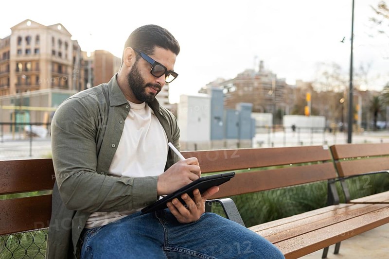 A man wearing sunglasses sits on a park bench, deeply focused on sketching on a digital tablet with a stylus. Urban buildings form the backdrop, creating a serene atmosphere in the city