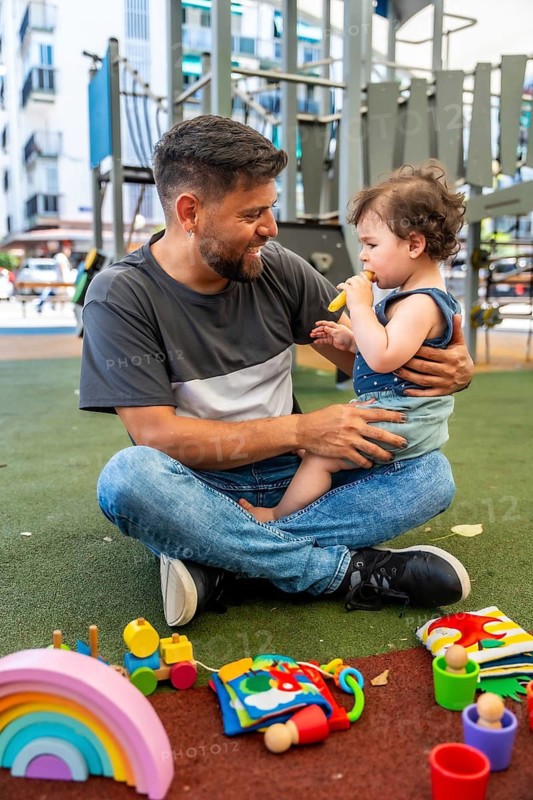 Happy dad sitting criss cross applesauce on the soft ground, embracing his toddler son who is holding a banana, surrounded by colorful children's toys on a sunny day at the playground