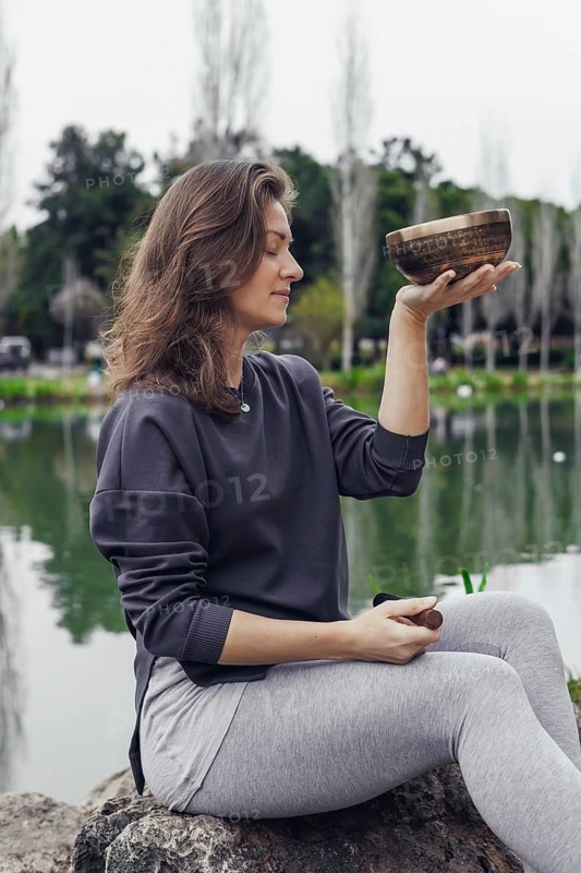 A young Caucasian woman with brown hair sits by a lake, holding a bowl. She wears a gray sweater and light gray pants. The background features trees and water