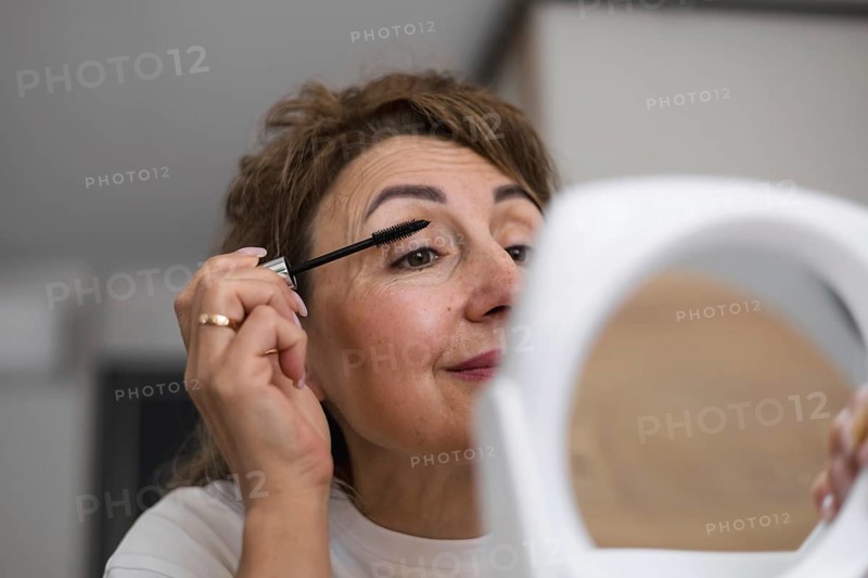 Mid aged woman prepares for her day by applying mascara while sitting at a wooden table in a bright, cozy room. The mirror reflects her focused expression as she enhances her beauty. Morning routine