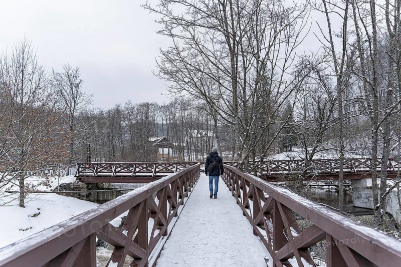 A man crosses the river in winter on a wooden bridge, A person walks alone on a snow-covered wooden bridge in a winter woodland scene, Belmontas park, Vilnius, Lithuania