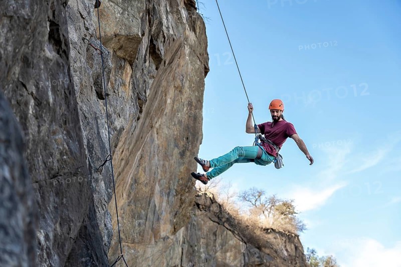 A determined young man ascends a steep cliff, using a rope for support, highlighting his strength and focus as he navigates the challenging rock face, embodying the essence of adventure and perseverance