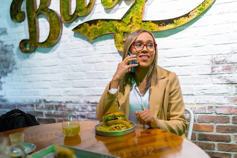 Latin woman talking to the mobile while eating burger in a modern fast food restaurant
