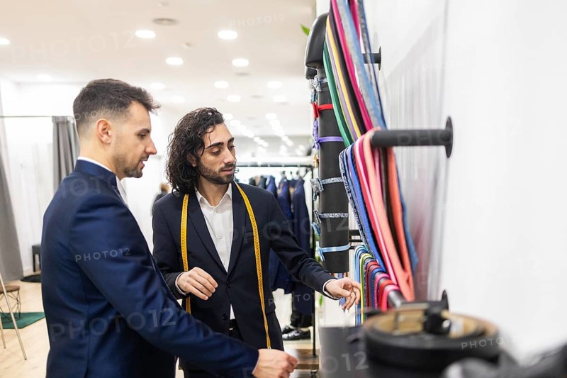 A tailor helps a client choose a suit in a suit store with colorful ties. Both are dressed in formal attire, highlighting fashion and style