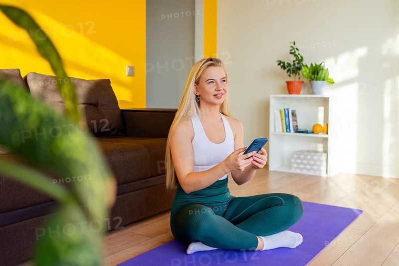 A woman in active wear sits cross legged on a yoga mat in a cozy home. She holds a smartphone, surrounded by modern decor and a vibrant yellow accent wall