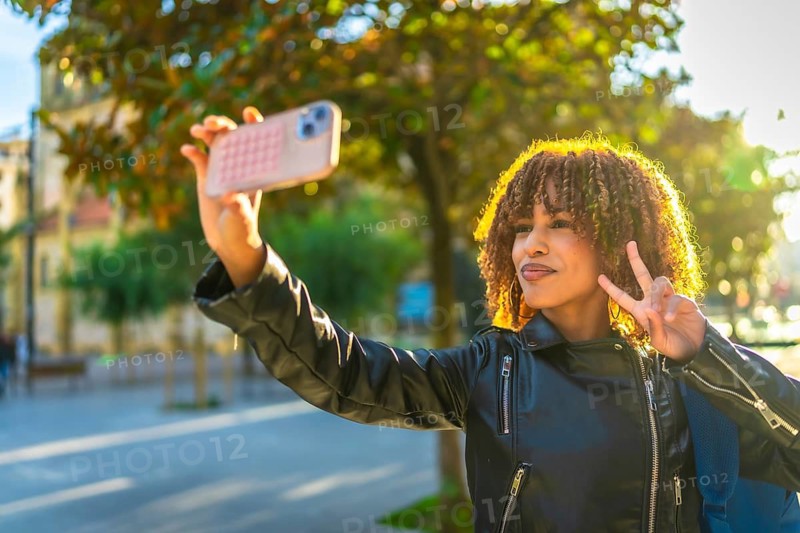 Beauty latin woman with curly hair gesturing success while taking selfie in the city during sunset