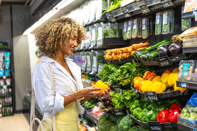 Woman with curly hair smiling while choosing a fresh yellow bell pepper from a vibrant produce display, emphasizing healthy eating, urban lifestyle, and grocery shopping experience