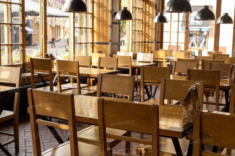 The interior of a cozy cafe features rows of simple wooden chairs and tables bathed in warm, natural light from large windows. The absence of customers creates a peaceful atmosphere, while the sunlight casts enchanting shadows on the floor. The inviting design highlights an ideal spot for relaxation or casual meetings