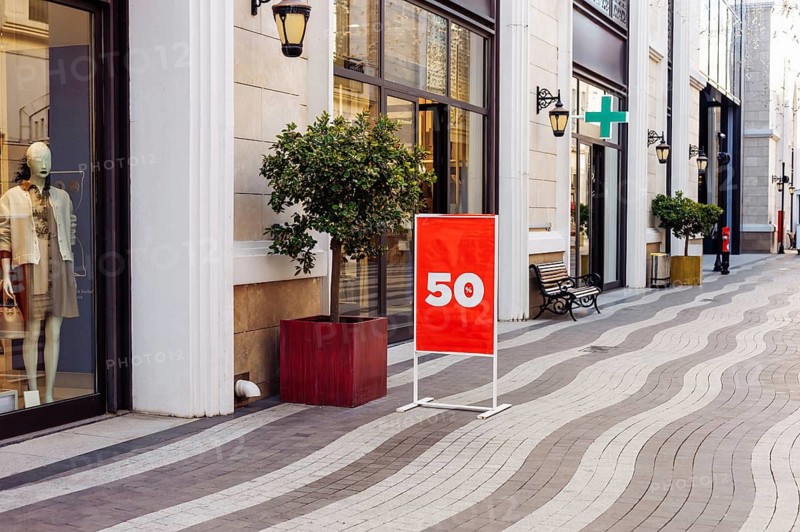 Mockup, retail storefront features bright red sign placed on clean street. Large windows showcase stylish clothing, while cheerful tree and bench encourage relaxation in this vibrant shopping area