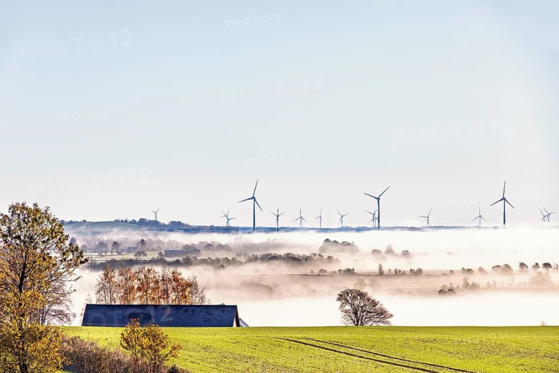 Wind turbines emerges above morning fog stretching across the countryside. Rolling farmland, scattered trees, and soft autumn colors create a peaceful rural landscape, Sweden