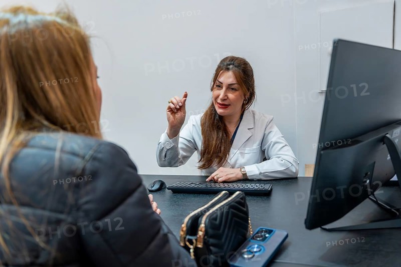 Female professional in a white coat consulting a young woman about a treatment, seated at a dark desk with a laptop, keyboard, mouse, wallet, and smartphone