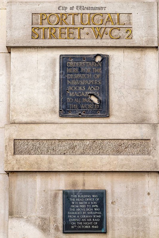 Two bronze plaques on Portugal Street mark the former head office of W H Smith and Son, which occupied the building from 1920 to 1976. A lower plaque records that shrapnel damage on the upper sign came from a German bomb during an air raid on 10 October 1940. London, England, United Kingdom