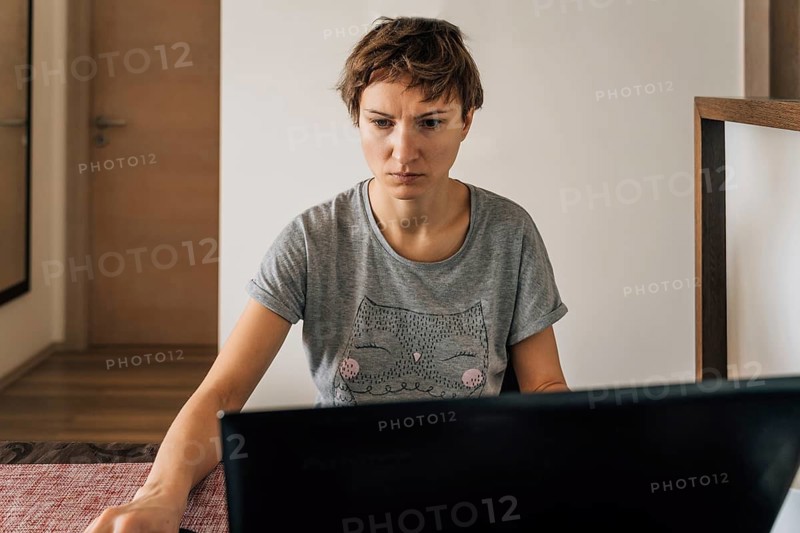 A woman in home clothes works at home at laptops with a tense look, A woman with short hair sits indoors concentrating on her work at a laptop