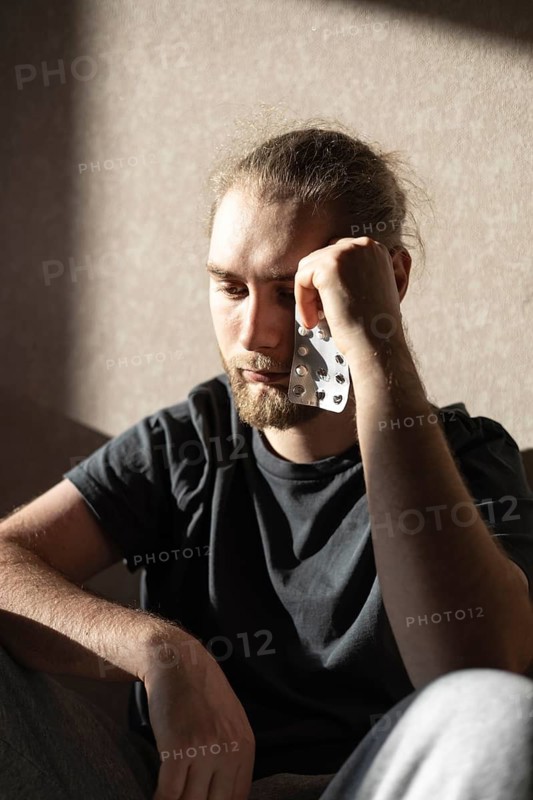 Young man with a beard thoughtfully holding a blister pack of pills. Sitting in a room, he appears deep in thought, suggesting themes of health or decision-making