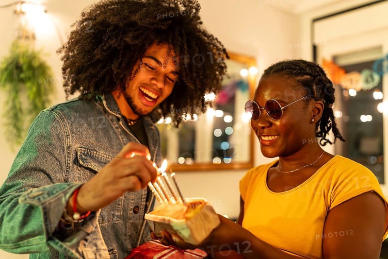 Multi-ethnic friends lighting birthday cake's candles preparing a surprise at home during party at night