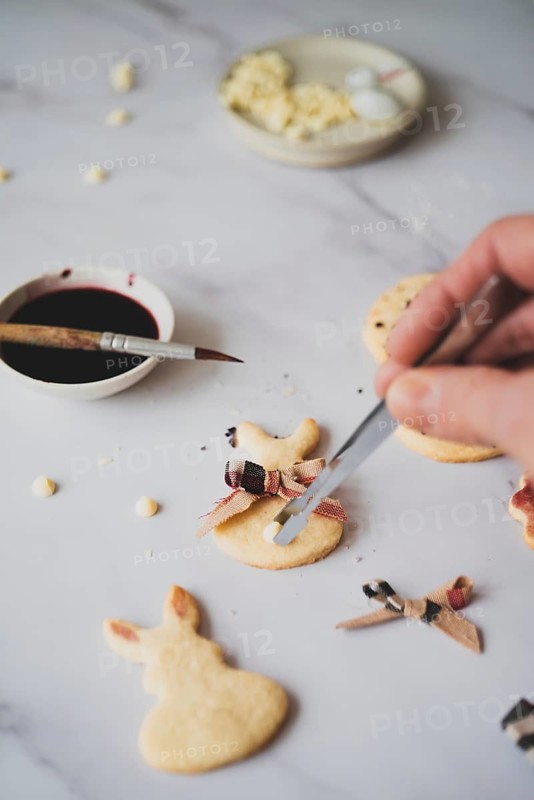 From above, a cropped unrecognizable person delicately paints Easter bunny shaped cookies with hibiscus tea using a thin brush, set beside a bowl of vibrant, dark red tea and scraps of dough