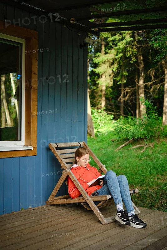 Young woman sits on wooden chair on terrace of tiny house and reading book. Trees and greenery surround her. It is daytime and the sun shines on the scene. She appears focused on her reading
