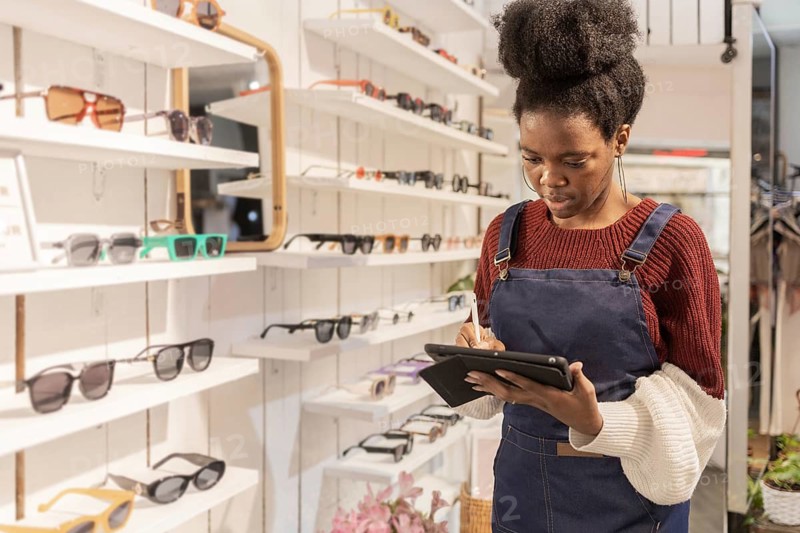 A woman in a fashionable store, surrounded by diverse sunglasses displayed on shelves. She is holding a tablet, likely checking inventory or browsing designs