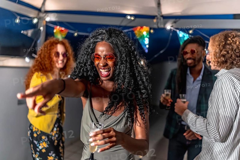 African woman wearing stylish night dress and sunglasses pointing ahead dancing with friends at night party in a terrace