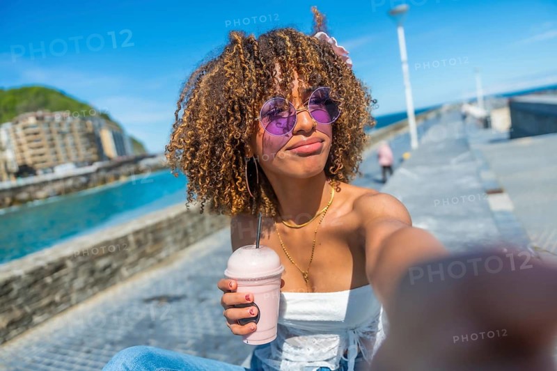 Young woman with curly hair drinking shake and taking selfie in the promenade next to the sea