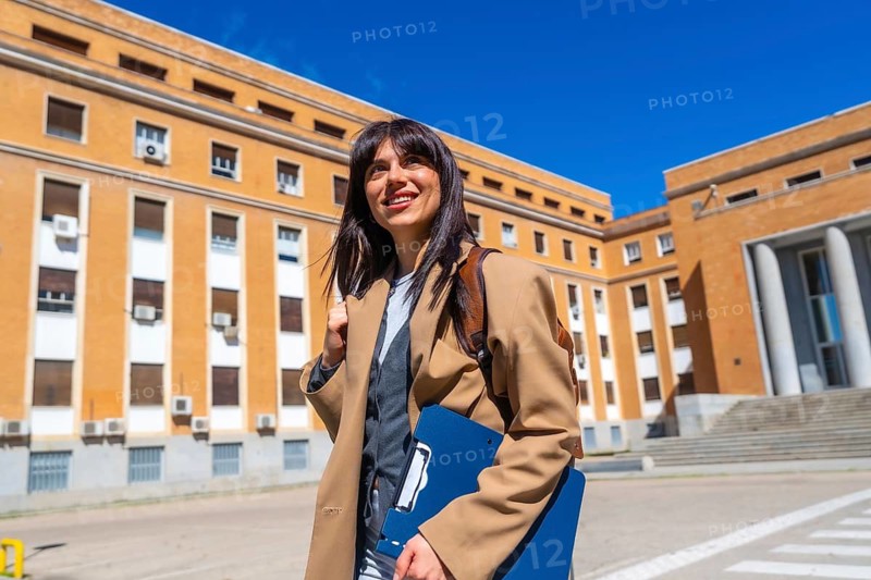 Young woman with a backpack and a blue clipboard walking confidently towards a university building, representing education, learning, and future success