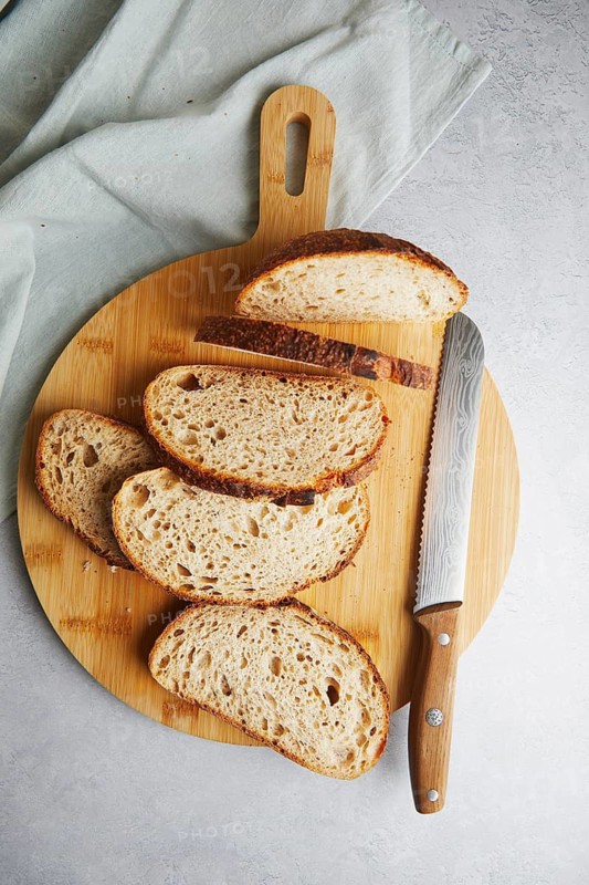 Top view of fresh homemade sourdough bread slices neatly placed on a wooden cutting board next to a stainless steel bread knife, with light gray textured background and a cloth partly visible