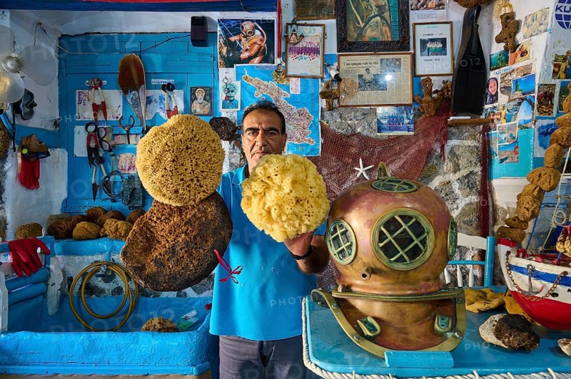 A man holds sponges next to an antique diving helmet in a blue, maritime-decorated room, Sponge Maufaktur, Nikolas Gourlas, Pothia, main town, port, Kalymnos, Sponge Diving Island, Dodecanese, Greek Islands, Greece