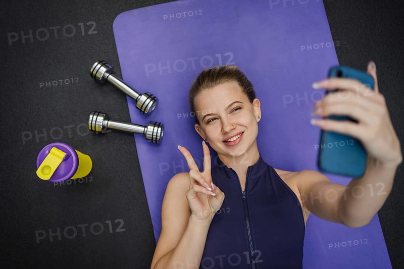 A blonde woman lies on a purple yoga mat, taking a selfie with a smartphone. Next to her are two dumbbells and a colorful water bottle, capturing a post workout moment