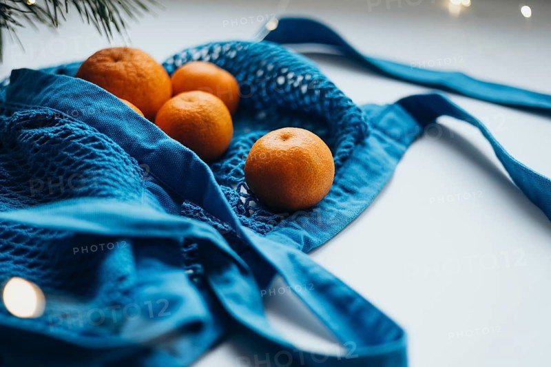 A vibrant blue string bag filled with juicy tangerines, elegantly positioned on a light table surface with subtle festive lights
