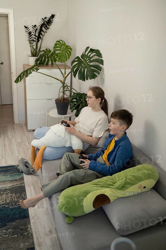 Siblings immerse in video game fun on a comfortable couch in a stylish living room, surrounded by plush toys and vibrant plants for a cozy ambiance
