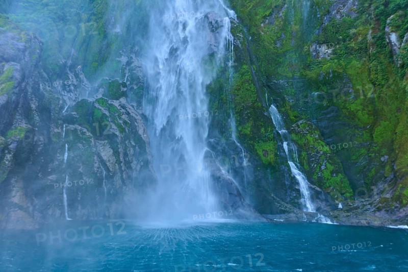 Waterfall pours over overgrown cliffs into the blue water, summer, Milford Sound, Te Anau, Fiordland National Park, South Island, New Zealand