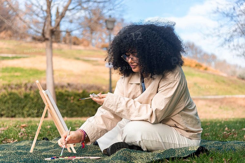 A young black woman with curly hair focuses on painting in a serene park, surrounded by vibrant green grass and trees, capturing creativity and relaxation in an outdoor environment