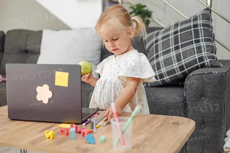 A young Caucasian girl with blonde hair stands by a laptop on a wooden table. Colorful toys and stationery are scattered around her in a cozy living room setting