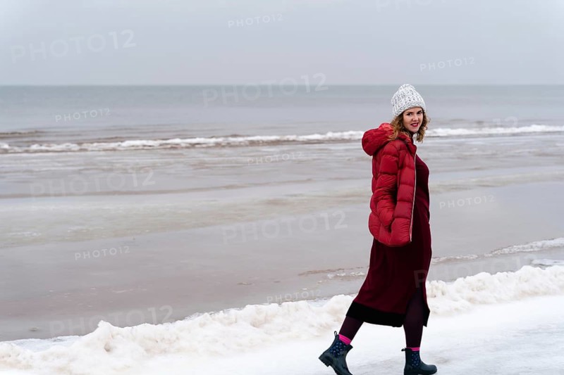 A woman in a red jacket and a red knitted dress walks in winter along the sea, A woman in a red coat and winter hat walks along a snowy beach with overcast skies, Jurmala, Latvia