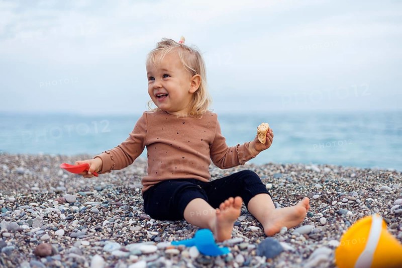 A young child sits on a pebble beach, holding a snack in one hand. The child girl appears curious and focused, with the sea stretching out in the background under clear sky