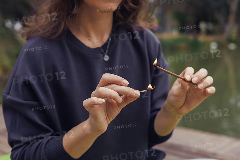 A young Caucasian woman with long brown hair sits by a pond, holding an incense stick. Smoke rises gently from the stick, creating a serene atmosphere