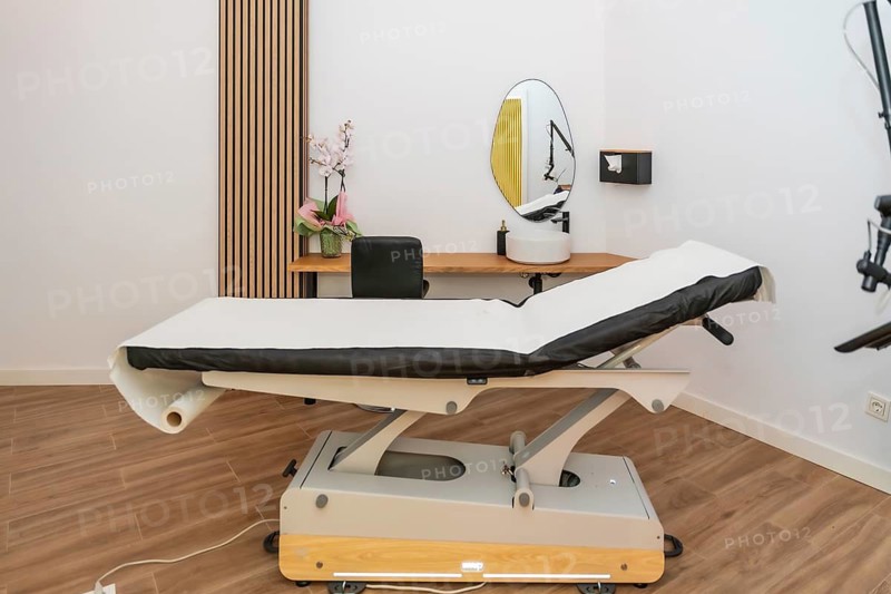 Clinic treatment room with adjustable medical bed covered in disposable paper, laser hair removal device, mirror and washbasin on wooden shelf, bright modern sterile interior