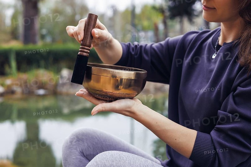 A young woman with brown hair plays a singing bowl outdoors by a serene body of water. She wears a dark sweater and gray leggings, promoting relaxation and mindfulness