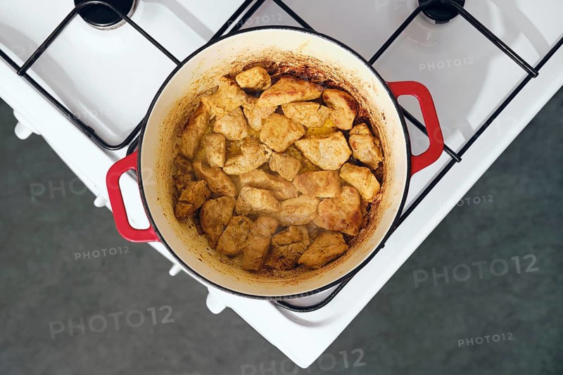 Pork pieces are cooking in red cast iron pot on white gas stove. Pot has brown crust on meat. Steam rises from pot. It is daytime in home kitchen setting