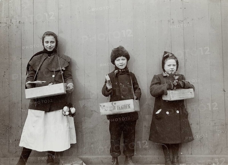 Berlin, Potsdamer Platz, Christmas, children selling toys, child labor, poverty, Three children wearing historic clothes and holding sales boxes in front of a wooden wall, 1908