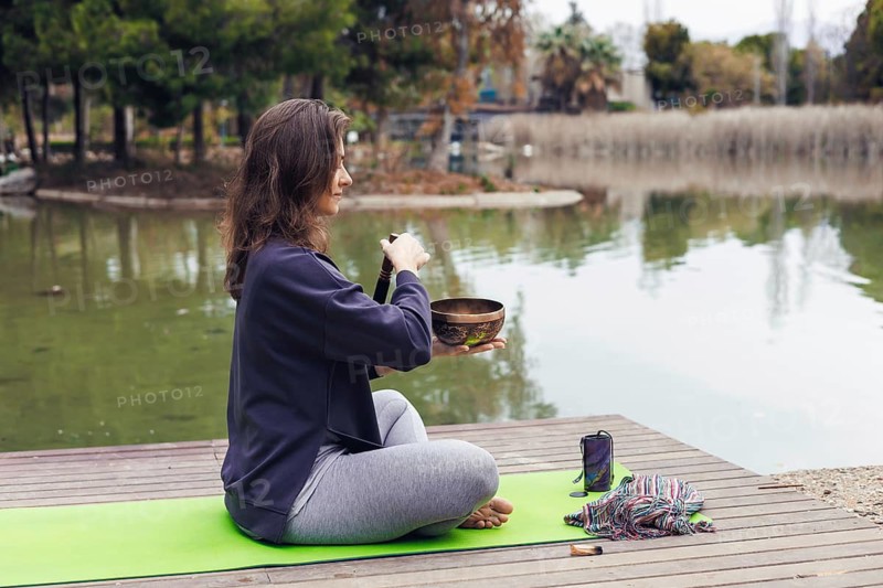 A young Caucasian woman with long brown hair sits cross-legged on a green yoga mat by a lake. She holds a singing bowl and appears to be meditating