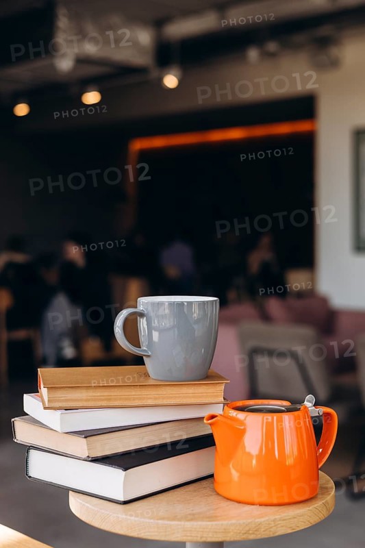 A cozy coffee shop scene with a grey mug placed on a stack of books, accompanied by an orange teapot on a wooden table. The background is softly blurred, creating a warm ambiance