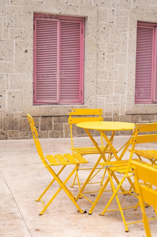 A cozy outdoor setting features vibrant yellow chairs and a table against a textured stone wall with charming pink shutters. The scene captures a relaxed and inviting atmosphere. Positive atmosphere