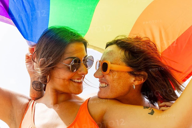 A joyful lesbian couple embraces under a vibrant rainbow flag, radiating happiness and connection on a sunny day. Their smiles and sunglasses add a playful vibe