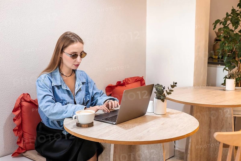 A young Caucasian woman with long blonde hair works on a laptop at a cafe table. She wears sunglasses and has a cup of coffee beside her